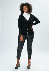 Cardigan nero con scollatura a V e chiusura a bottone, abbinato a pantaloni in pelle nera. Il modello indossa un dolcevita bianco e scarpe con tacco spesso.