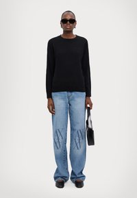 Maglione nero a maglia con scollo rotondo, jeans in denim azzurro chiaro con loghi in rilievo, borsa nera e scarpe slide nere.