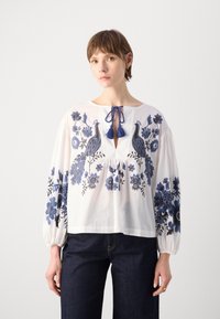 Weißes Blusenshirt mit blauen Blumen- und Pfau-Stickereien, Schlüsselloch-Ausschnitt mit blauer Bindung und Quasten sowie puffierten langen Ärmeln.