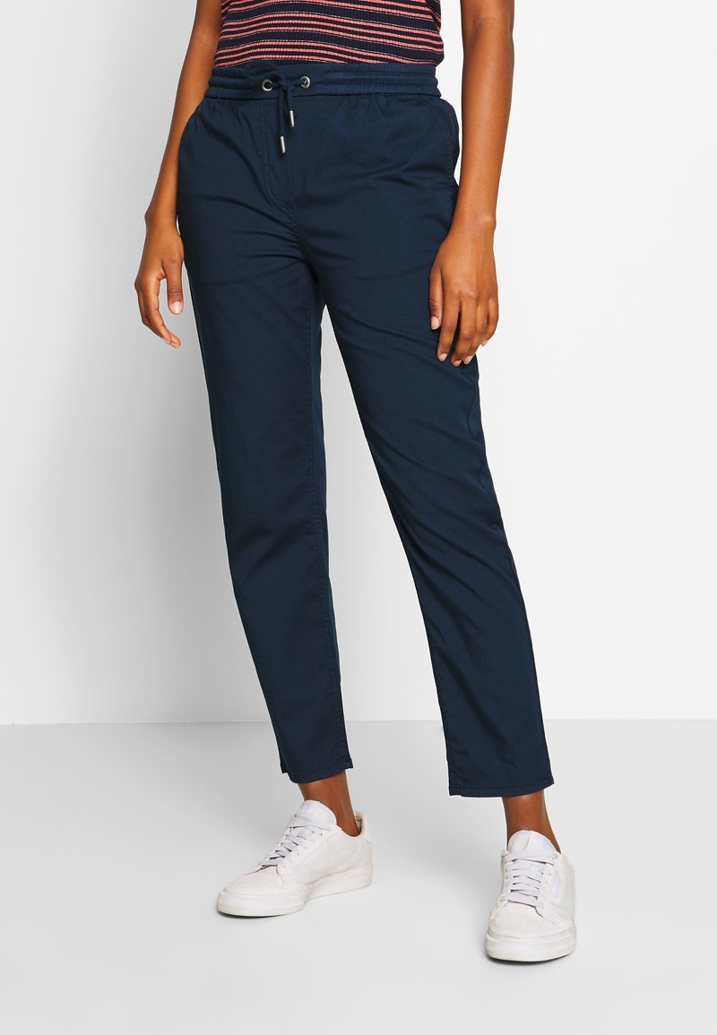 Personne portant un pantalon bleu marine à cordon, des baskets blanches et un haut rayé sur un fond uni clair.