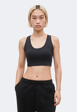 Νέα γυναίκα με κοντά ξανθά μαλλιά που φοράει μαύρο αθλητικό σουτιέν και μαύρο παντελόνι φόρμας, με τα χέρια στις τσέπες, στέκεται μπροστά σε λευκό φόντο.