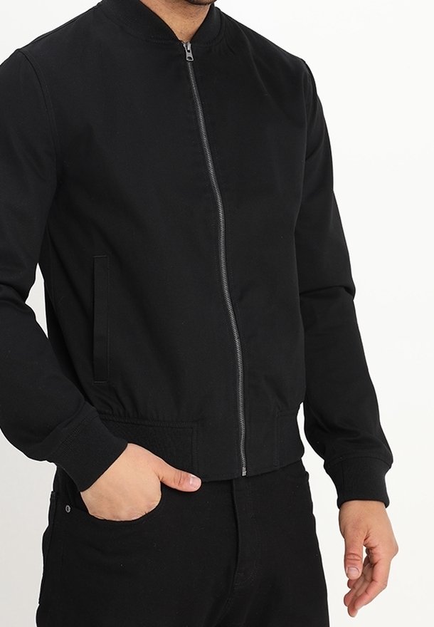 Černá bomber bunda vyrobená z měkké tkaniny, s stojáčkovým límcem, předním zipem a bočními kapsami. Detail žebrovaných manžet a spodního lemu.