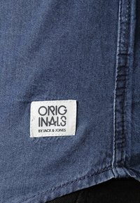 Sininen farkkukangas, jossa on ommeltu etiketti, jossa lukee "ORIGINALS" ja "BY JACK & JONES" paksulla mustalla tekstillä valkoisella taustalla.