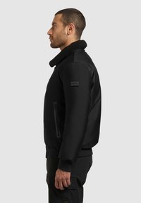 Jachetă bomber neagră cu guler texturat din pluș, buzunar laterat cu fermoar, manșete elastice și finisaj din țesătură moale. Include o patch cu logo pe mânecă.