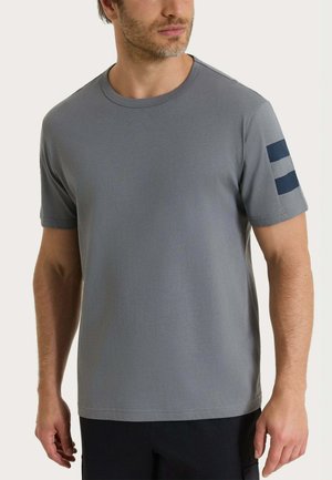 Uomo che indossa una maglietta grigia semplice a maniche corte con due strisce blu scuro sulla manica sinistra, abbinata a pantaloni neri.