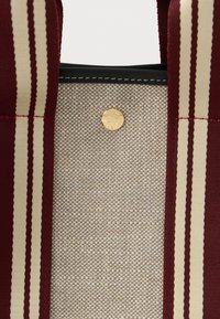 Κοντινό πλάνο από υφασμάτινη επιφάνεια σε μπεζ χρώμα μεταξύ δύο ριγέ λωρίδων σε μπορντό και κρέμα με ένα μικρό, στρογγυλό, χρυσό κουμπί.