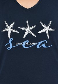 Tre stelle marine bianche sopra il testo che dice "HO BISOGNO DI VITAMINA mare" in bianco e blu su una maglietta blu navy a V.