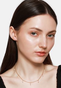 Goldene Halskette mit kleinen, tropfenförmigen Perlen, die daran h�ängen, getragen von einem Modell mit glattem, dunklem Haar und klarer Haut.