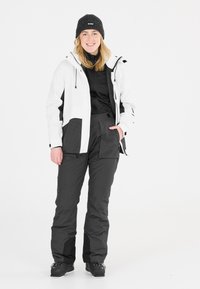 Witte en zwarte winterjacket met capuchon en ritszakken, gecombineerd met zwarte broek. De outfit omvat een donkere gebreide beanie en skibotsen.