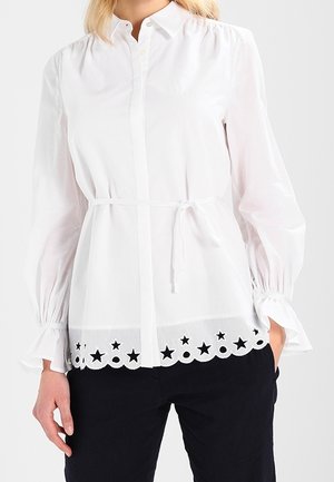 Hvid bluse med lange ærmer, flæsekant ved manchetterne, knapper foran, bælte i taljen og stjerneformet udskæringsmønster ved kanten, kombineret med sorte bukser.