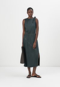 Denim midi dress in dark wash with a side slit and asymmetrical seams. Features a one-shoulder design with a bow detail and casual sandals.