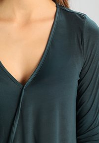 Türkisfarbener V-Ausschnitt mit langen Ärmeln aus glattem Stoff, mit einer vorteilhaften Drapierung und Nahtdetails entlang des Ausschnitts.