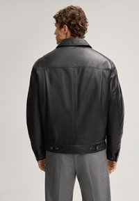 Giacca in pelle nera con colletto ampio, dettagli cuciti e bottoni laterali. Texture liscia con vestibilità comoda, indossata sopra pantaloni grigi.
