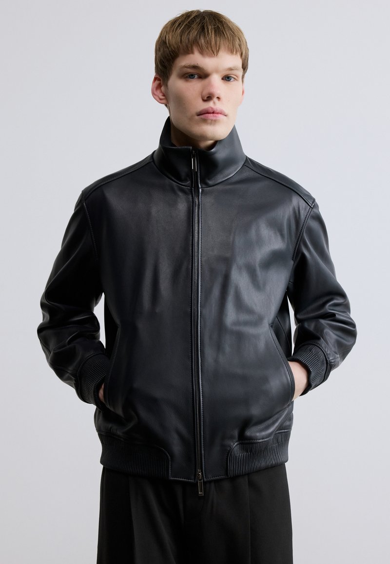 Fekete bőr bombázódzseki magas gallérral, cipzáras záródással és bordázott mandzsettákkal. Sima textúra és minimalista dizájn.