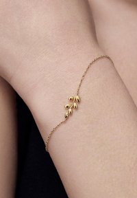 Złota bransoletka z delikatnym wzorem liścia, z polerowanymi akcentami liści na cienkim łańcuszku. Noszona na jasnej karnacji, w minimalistycznym stylu.