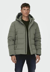 Lichtgroene pufferjack met capuchon, met een ritssluiting aan de voorkant, drukknoopsluiting en een gewatteerde textuur. G gedragen over een donkergroene trui.