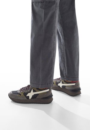 Gambe che indossano pantaloni grigi e sneaker multicolori con design a stelle bianche e suole scure macchiate, su uno sfondo bianco.