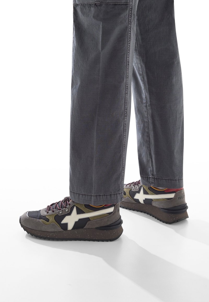 Ben klädda i grå byxor och flerfärgade sneakers med vit stjärndesign och mörka fläckiga sula, mot en vit bakgrund.