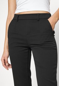 Pantalons noirs ajustés en tissu lisse, dotés de passants de ceinture, de poches latérales et de plis pour une apparence structurée.