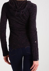 Person, der ein dunkel gehäuften Langarmoberteil mit subtilen rosa Streifen und eine dunkelblaue Jeans trägt, von hinten gezeigt.