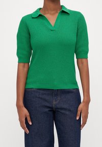 Grön stickad pikétröja med korta ärmar, med ribbad struktur och V-ringad krage. Kombinerad med mörk denimjeans.