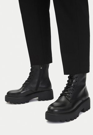 Botas de tobillo de cuero negro con cordones y suelas gruesas con tacos, y cremalleras laterales, combinadas con pantalones negros sobre fondo blanco.