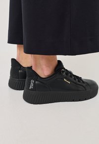 Melnas kedas ar teksturētu ādu un gludu virsmu, ar rievotu zoli, zelta akcentiem un perforētu papēža detaļu.