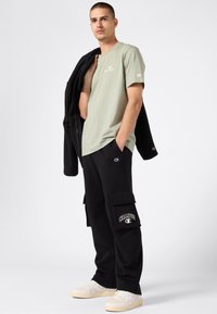 Camiseta verde claro con logo, pantalones cargo negros con bolsillos y zapatillas color crema. El modelo lleva una chaqueta negra texturizada sobre la camiseta.