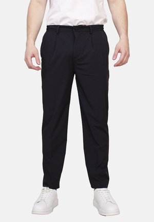 Uomo che indossa pantaloni neri su misura, sneaker bianche e una camicia bianca, in piedi davanti a uno sfondo uniforme.