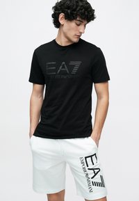 Νέος άνδρας που φοράει μαύρο t-shirt EA7 Emporio Armani και λευκά σορτς με λογότυπο EA7, με τα χέρια στις τσέπες, κοιτάζοντας προς τα κάτω πλάγια.