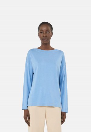 Donna in piedi di fronte, indossa un maglione azzurro chiaro a maniche lunghe e pantaloni beige su uno sfondo bianco semplice.