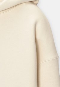 Sweat à capuche beige à manches longues avec des détails de coutures visibles sur la manche et la capuche, fabriqué en tissu doux et texturé.