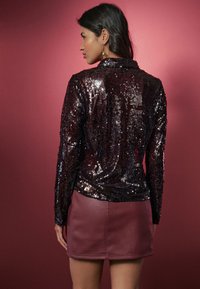 Pikkade varrukatega pluus, kaetud läikivate mustade litritega, kombineeritud keha järgiva burgundia nahkkleitiga. Kangas on sile ja läikiva väljanägemisega.