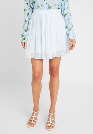 Kvinde iført hvid plisseret nederdel, blomstret bluse med lange ærmer og hvide sandaler med stropper og åbne tæer, stående mod en ensfarvet baggrund.