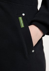 Main insérée dans la poche d'un sweat-shirt noir avec une étiquette en tissu verte et des détails de bouton en métal sur le bord de la poche.