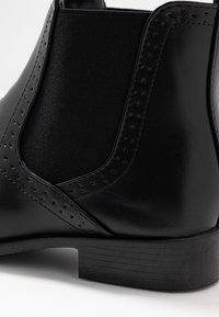 Stivaletto Chelsea in pelle nera con pannelli elastici laterali, dettagli traforati e tacco basso impilato. Texture liscia con un design elegante.