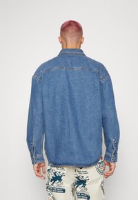 Denim πουκάμισο σε ανοιχτό μπλε απόχρωση με κοφτό γιακά, μακριά μανίκια, κουμπωτές μανσέτες και λεπτομέρειες στο γιακά. Διαθέτει ραμμένες ραφές και άνετη εφαρμογή.