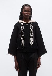 Sudadera con capucha negra con cremallera, detalles beige y cordones con estampado de cebra. Presenta un corte holgado y mangas anchas. Textura suave.