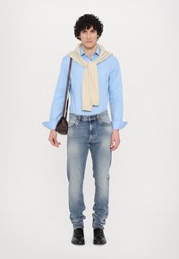 Lichtblauw longsleeve shirt met opgerolde mouwen, beige sweater over de schouders, vervaagde blauwe jeans en zwarte schoenen, terwijl een kleine bruine tas wordt vastgehouden.