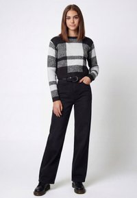 Sort-hvid ternet strikket sweater med lange ærmer, kombineret med sorte bukser med vide ben og sorte sko. Modellen står med hænderne i lommerne.