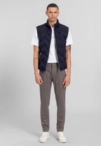 Marineblaues gesteppte Weste mit hohem Kragen, Reißverschluss, getragen über einem weißen T-Shirt, kombiniert mit grauen Jogginghosen und weißen Sneakers.