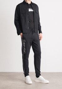 Black lightweight shirt with two chest pockets, paired with black joggers featuring white text and side stripes, completed with white sneakers.