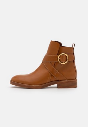 Bottine en cuir marron avec un petit talon en bois, des brides croisées décoratives et une boucle ronde dorée sur fond blanc.