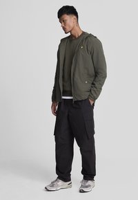 Giacca a zip verde oliva con cappuccio, con logo giallo; abbinata a pantaloni cargo scuri e sneakers grigio chiaro. Design semplice e funzionale.