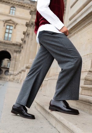 Pantaloni larghi grigi realizzati in tessuto morbido, abbinati a scarpe elegante in pelle lucida marrone scuro, inseriti in un contesto di architettura storica.