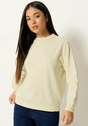 Jeune femme portant un sweat-shirt jaune pâle à manches longues avec des boutons sur l'épaule et un jean bleu foncé.
