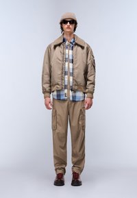 Beżowa kurtka bomberka z futrzanym kołnierzem, nałożona na niebieską i białą koszulę w kratę z guzikami. Beżowe spodnie cargo i ciemne buty z czerwonymi sznurówkami.