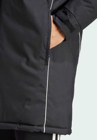 Schwarze isolierte Jacke mit weißen Paspeln, ausgestattet mit einer Seitentasche und einem Klettverschluss am Ärmel für eine verstellbare Passform. Glatte Textur.
