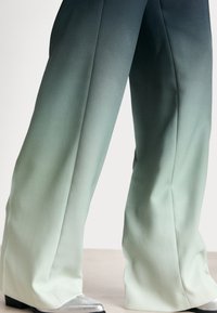 Weite Hosen mit einem Farbverlauf von dunkelgrün zu hellmint, mit einer glatten Textur und dezentem Glanz, kombiniert mit metallischen Schuhen.