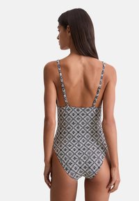 Femme avec de longs cheveux noirs portant un maillot de bain une pièce noir et blanc à motifs, vue de dos sur un fond uni.